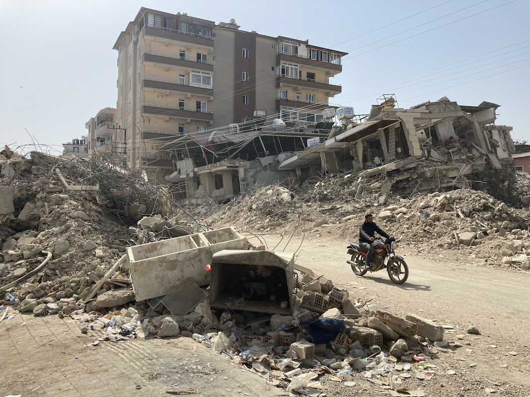 Foto: Ein Mann fährt auf einem Motorrad zwischen zerstörten Häusern hindurch.
