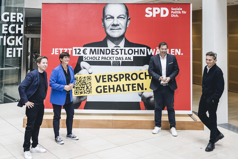 Foto: Die SPD-Parteispitze steht vor einem Mindestlohn-Plakat zur Bundestagswahl und klebt einen Störer "Versprochen. Gehalten."