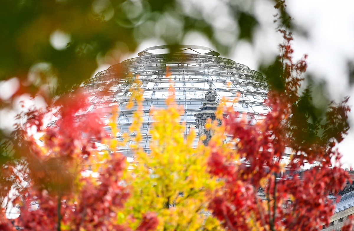 Foto: Rot-gelb-grün herbstlich verfärbte Blätter sind an Bäumen vor der Reichstagskuppel zu sehen.