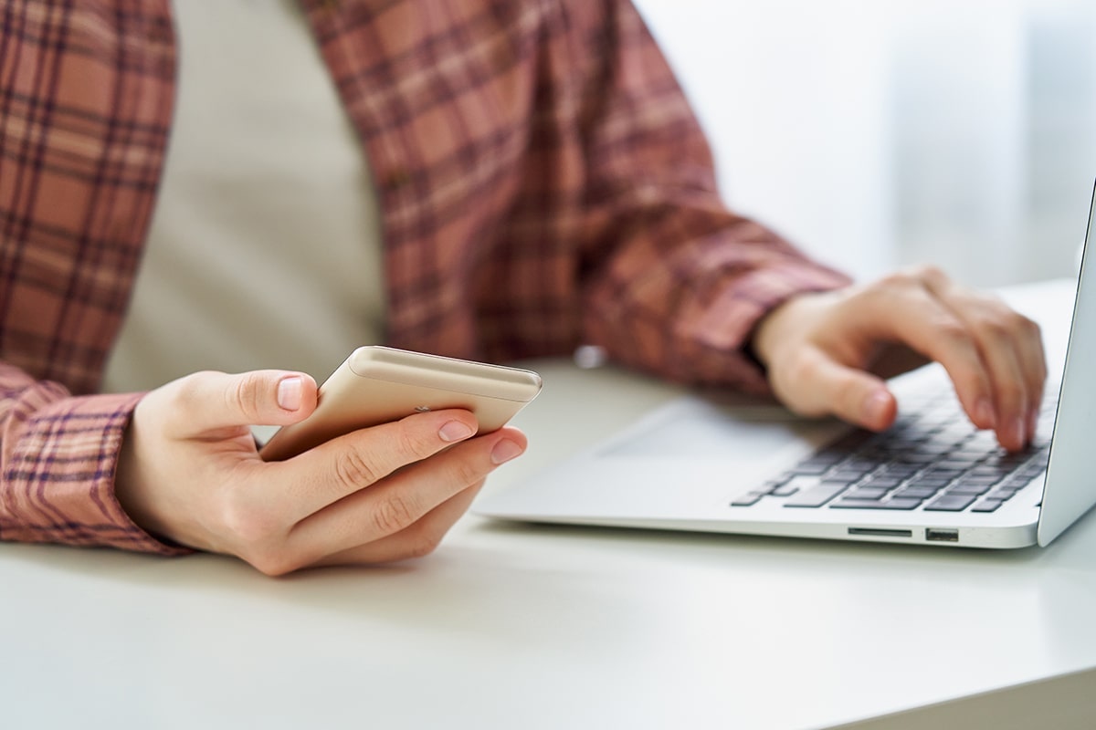 Foto: Gesichtslose junge Frau surft auf dem Smartphone während sie auf dem Laptop tippt