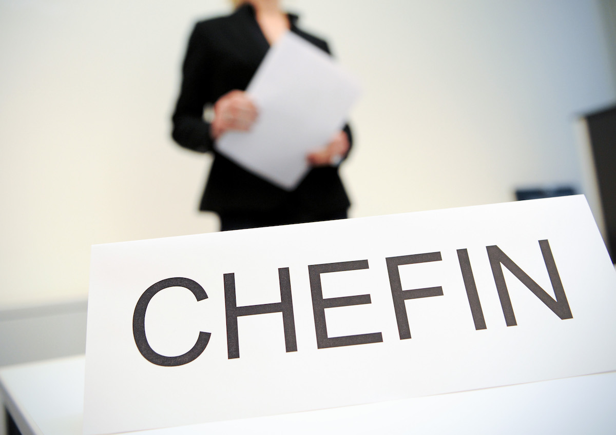 Foto: Eine Frau hält in einem Büro Papiere in der Hand. Vor ihr steht ein Schild mit der Aufschrift „Chefin“