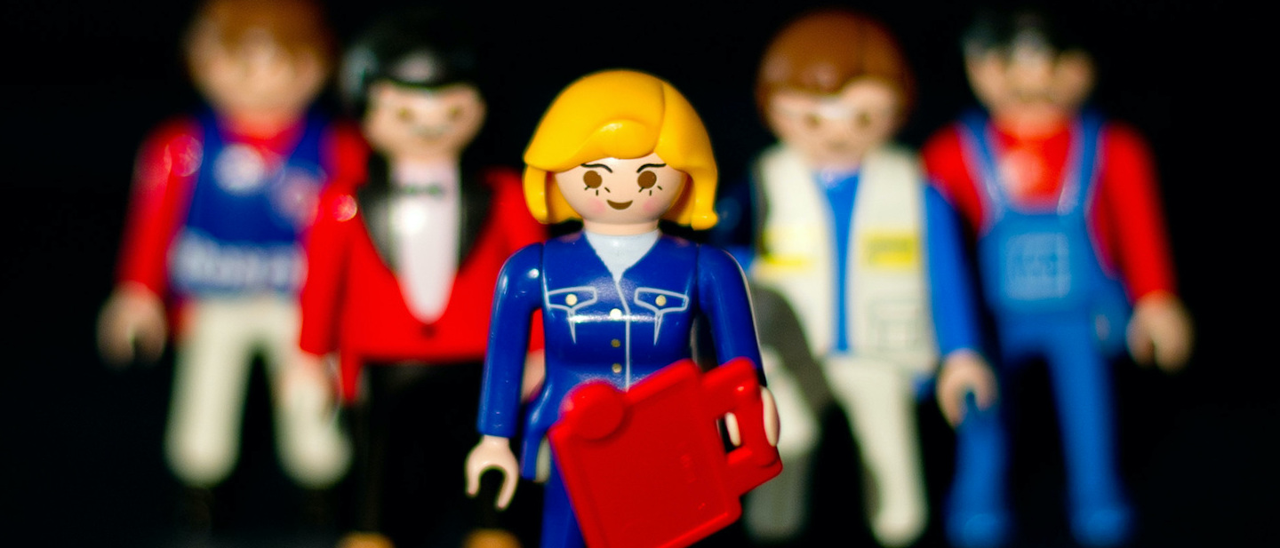 dpa Illustration zu Frauenquote: Eine weibliche Playmobil-Figur steht vor mehreren männlichen Figuren.