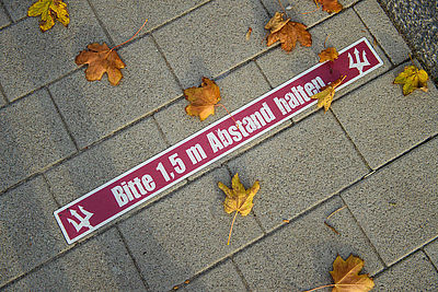 Foto: Herbstliche Blätter liegen neben einem Aufkleber mit der Aufschrift, "Bitte 1,5 m Abstand halten" auf dem Boden 