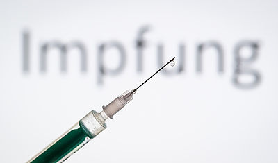 Foto: Eine Spritze wird vor den Schriftzug „Impfung“ gehalten. 