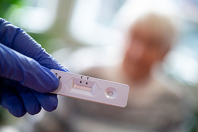 Foto: Altenpflegerin hält im Seniorenheim einen Antigen-Corona-Schnelltest in der Hand. Im Hintergrund sitzt eine Bewohnerin.